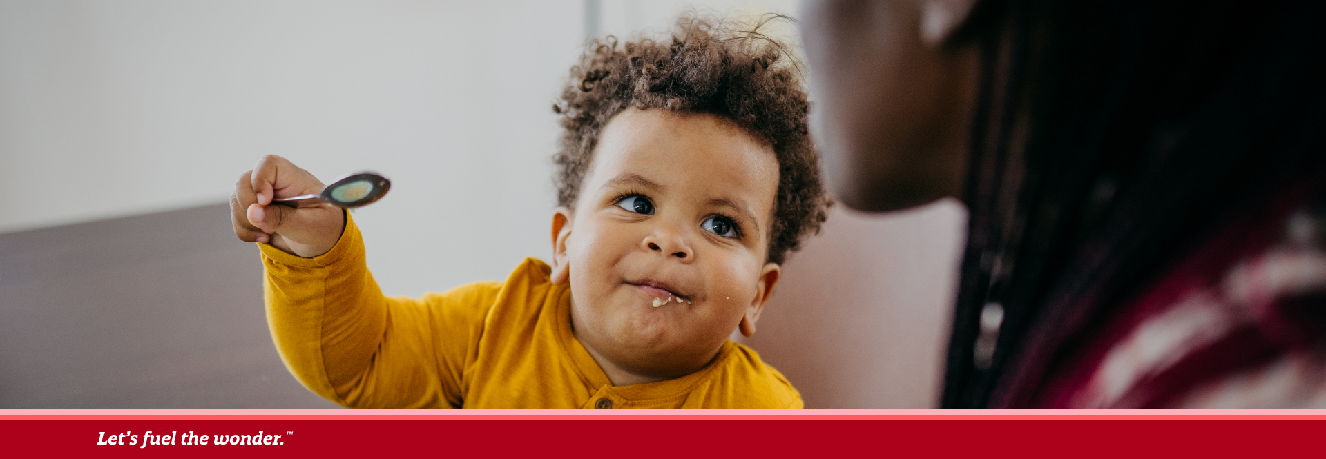 Toddler in a yellow shirt sitting at a table with a bowl of food, holding a spoon and looking up at an adult.