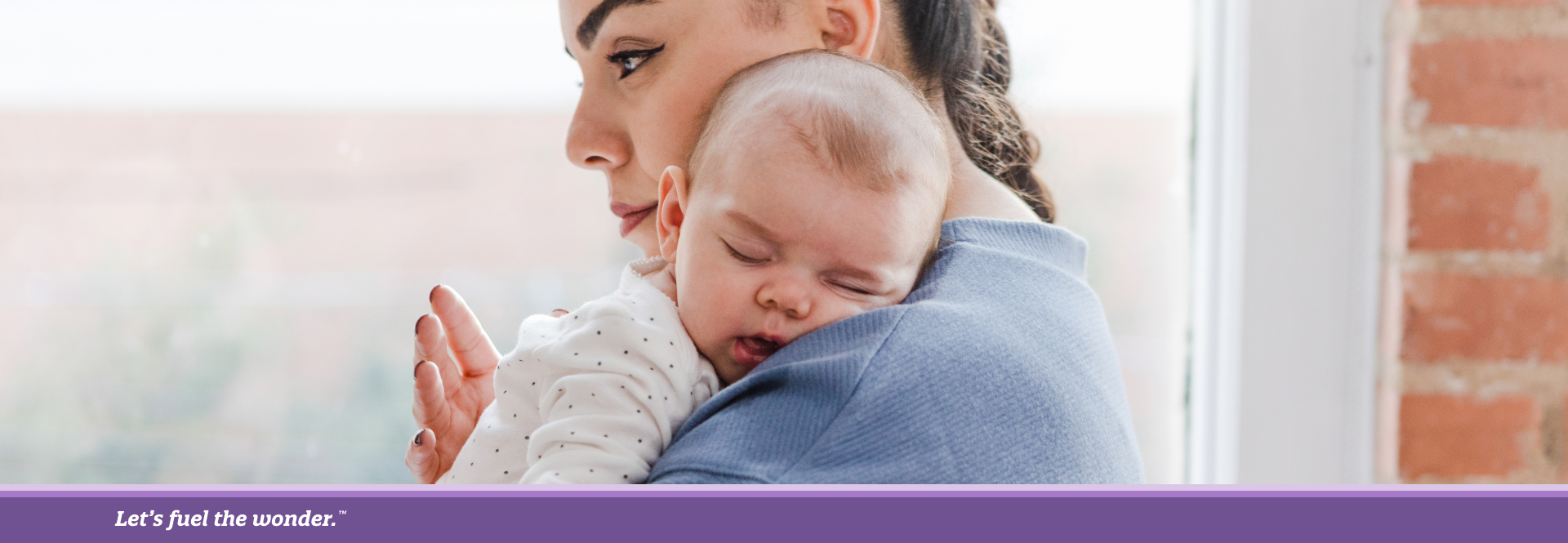 Woman holding a sleeping baby on her shoulder while standing by a window.