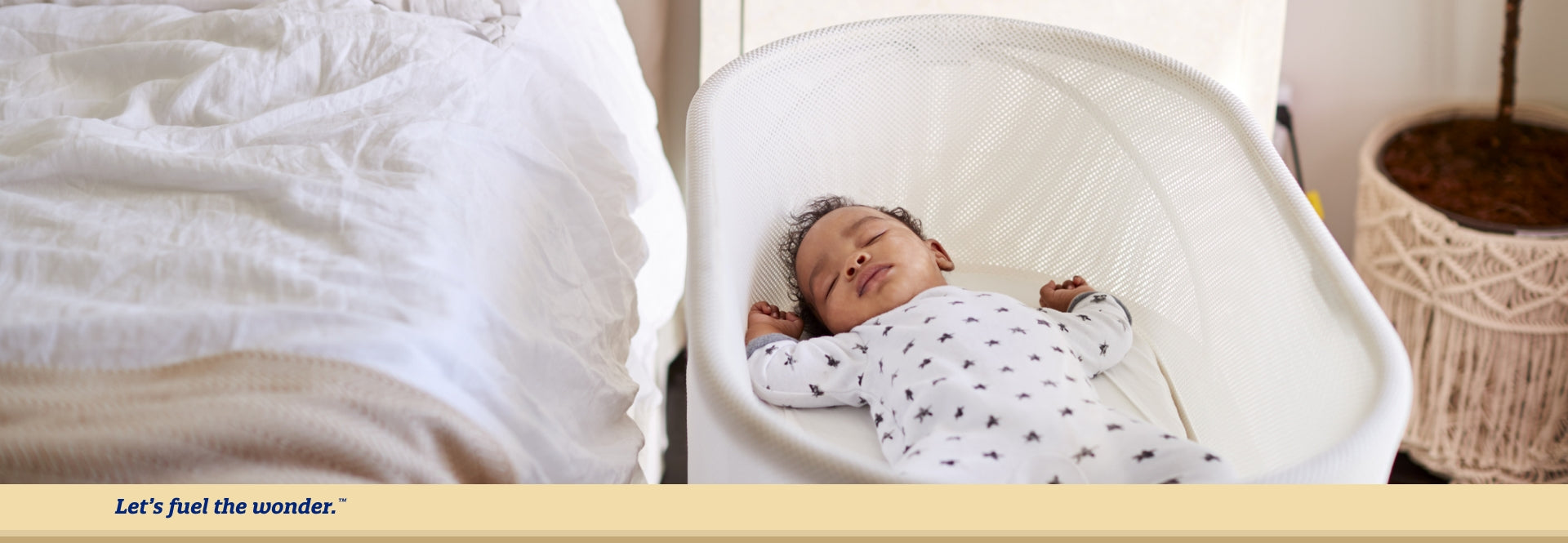 Sleeping baby laying on their back in bassinet next to larger bed in well-lit bedroom