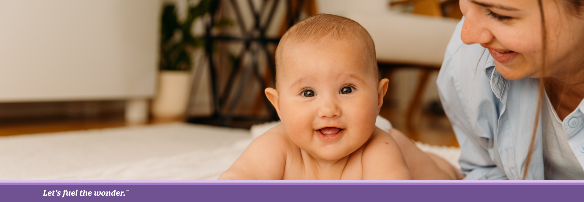 Smiling baby lying on stomach on a blanket while a caregiver looks on happily.