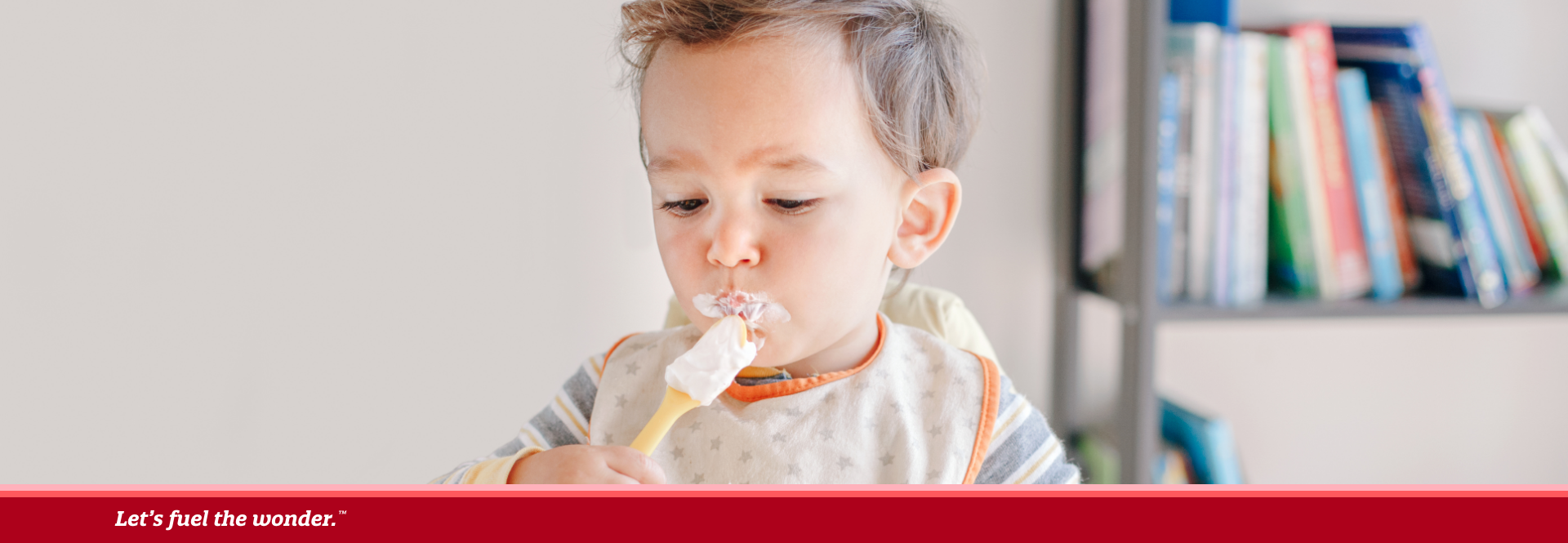 Toddler sitting in a high chair eating yogurt with fruit, holding a spoon and wearing a bib, with a sippy cup nearby.