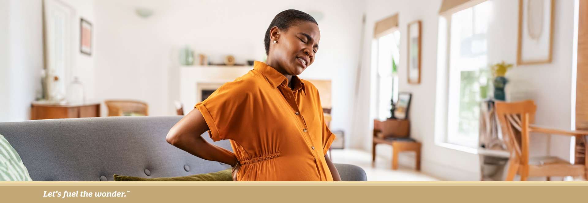 Pregnant woman in orange dress sitting on couch, holding her back and belly in discomfort.