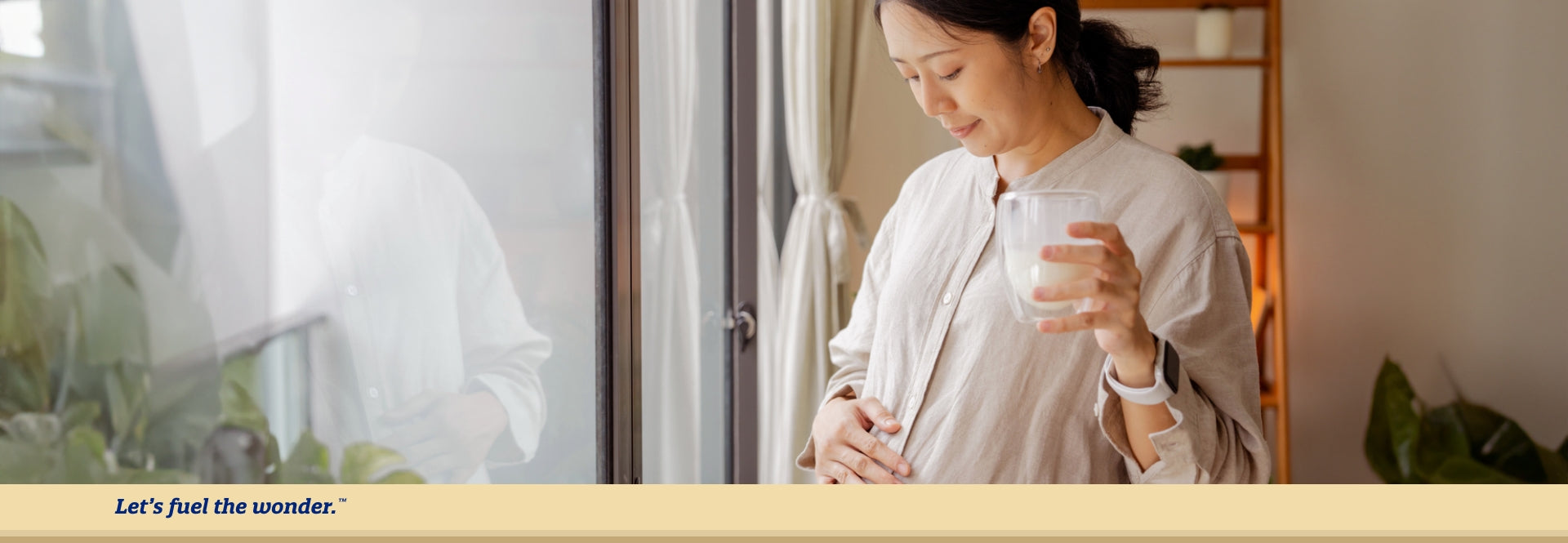 Pregnant woman standing by window looking down at bellow while holding a glass of milk in left hand and rubbing pregnant belly with right hand