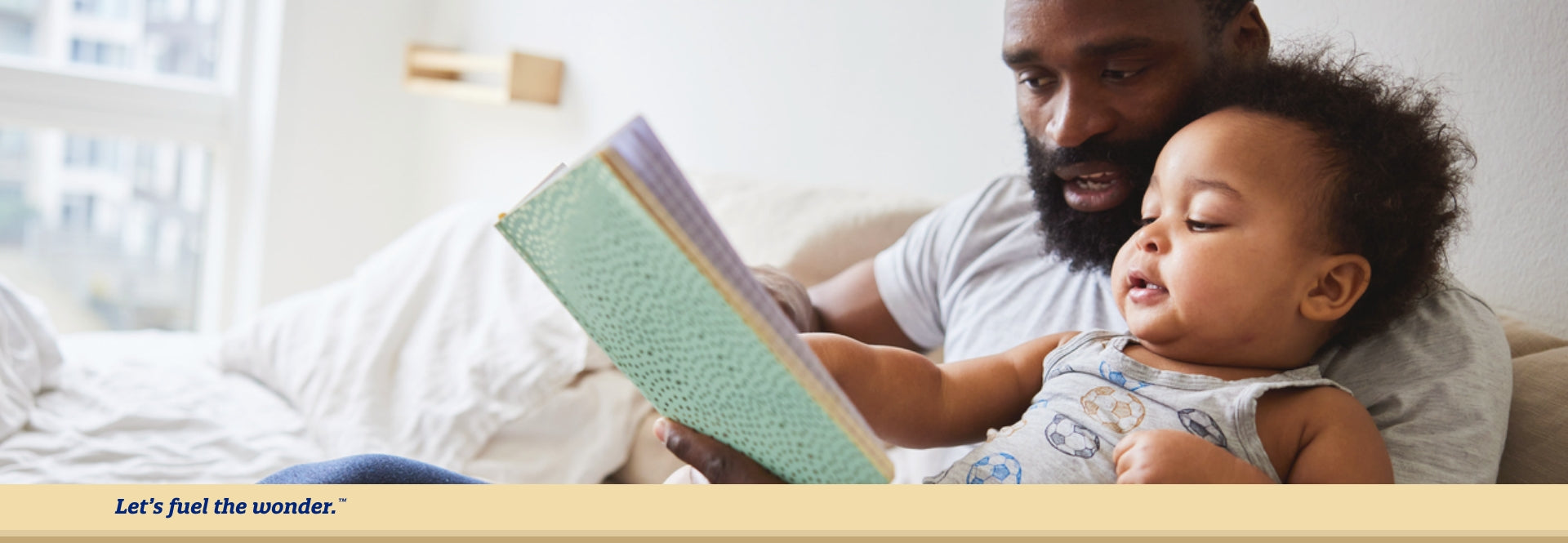 Young toddler and father sitting on a couch. Young toddler is sitting in father's left arm while pointing and reading a booklet held up by father.