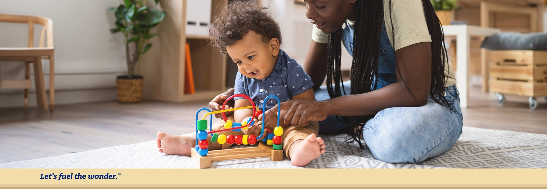 Young toddler sitting on ground, hunched over with legs stretched out while playing with a puzzle toy. Mother sitting on ground behind young toddler playing with the same puzzle toy.