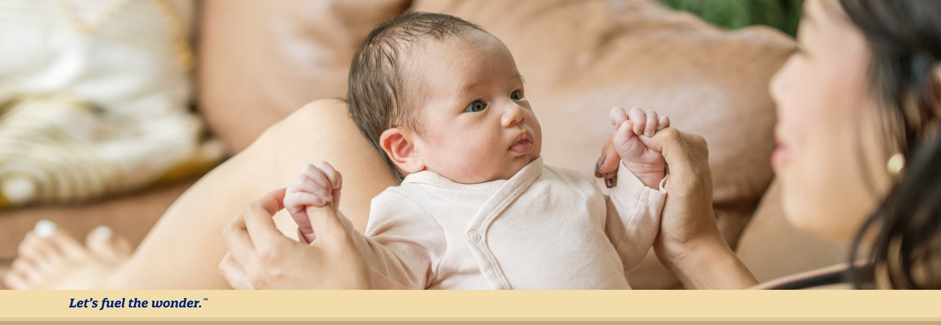 Baby with arms out staring at parent's face while baby is sitting upright on parent's legs. Parent is sitting up on couch while smiling at baby.