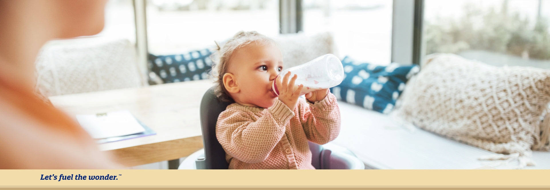 Young toddler sitting in high chair looking off to their left side as they are holding up and drinking from milk bottle