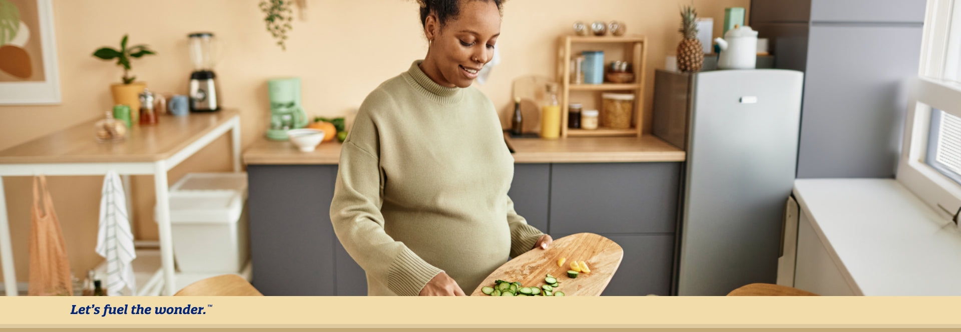Pregnant woman standing in kitchen looking down holding a cutting board with cucumber and lemon slices on top of cutting board.