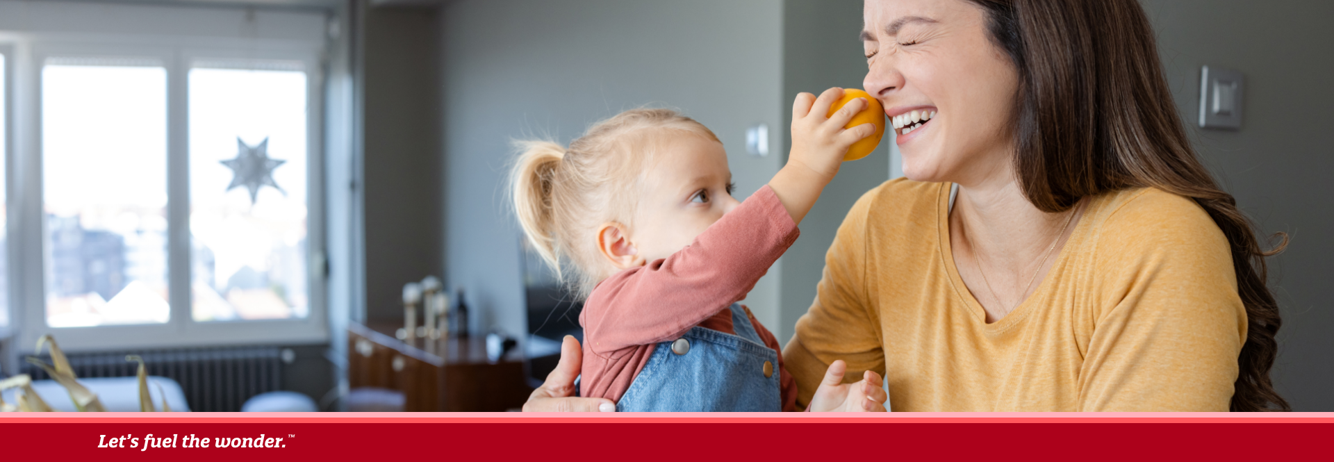 Smiling mother holding her young daughter, who playfully presses an orange to her mother's nose, with a basket of fresh fruits and vegetables in front of them.