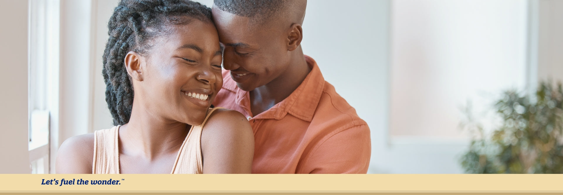 Smiling partner with eyes closed standing next to window as other smiling parent with eyes closed is hugging them from behind