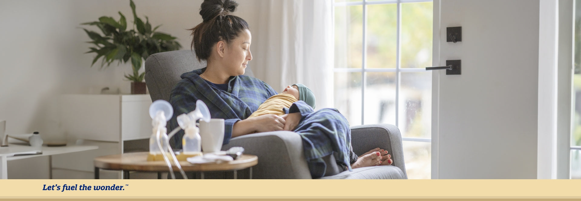 Mother sitting in single chair with legs crossed on cushion while holding sleeping newborn baby wrapped in a swaddle. A set of breast pumps are shown in the foreground.
