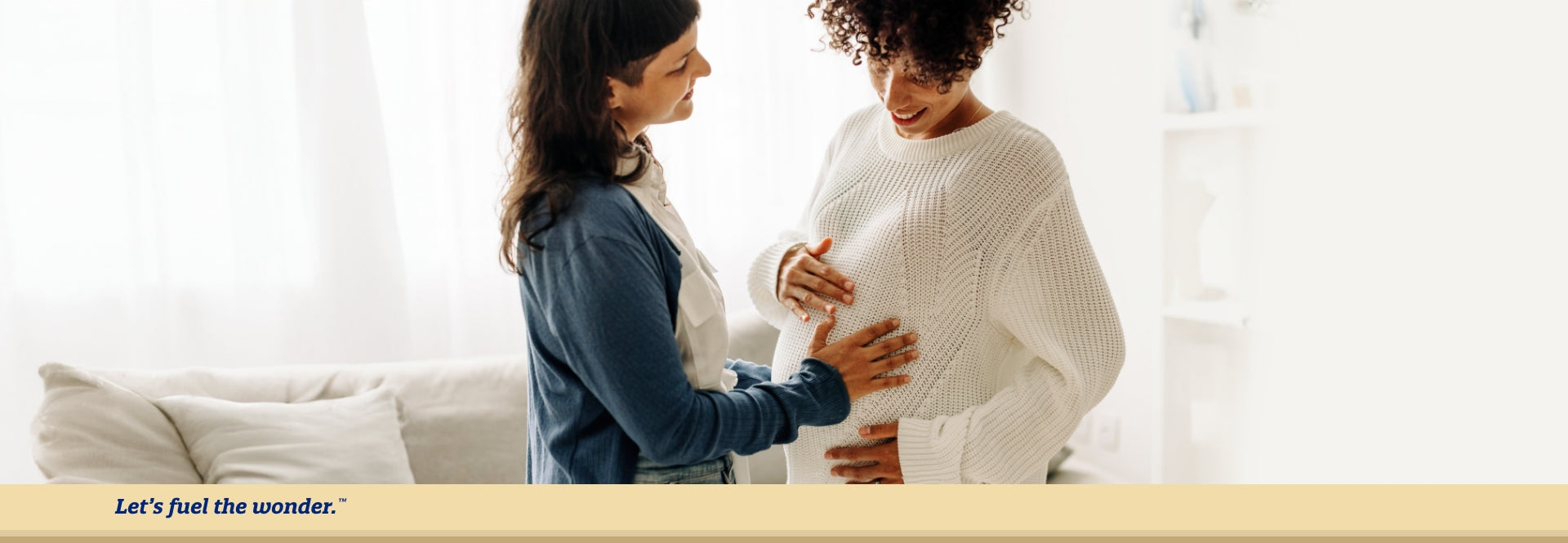 Pregnant parent smiling while standing in living room while smiling partner places their hand on pregnant parent's belly