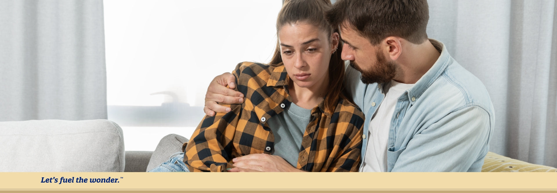 Upset partner sitting on couch while other partner sits next to them with arms wrapped around upset partner to console them