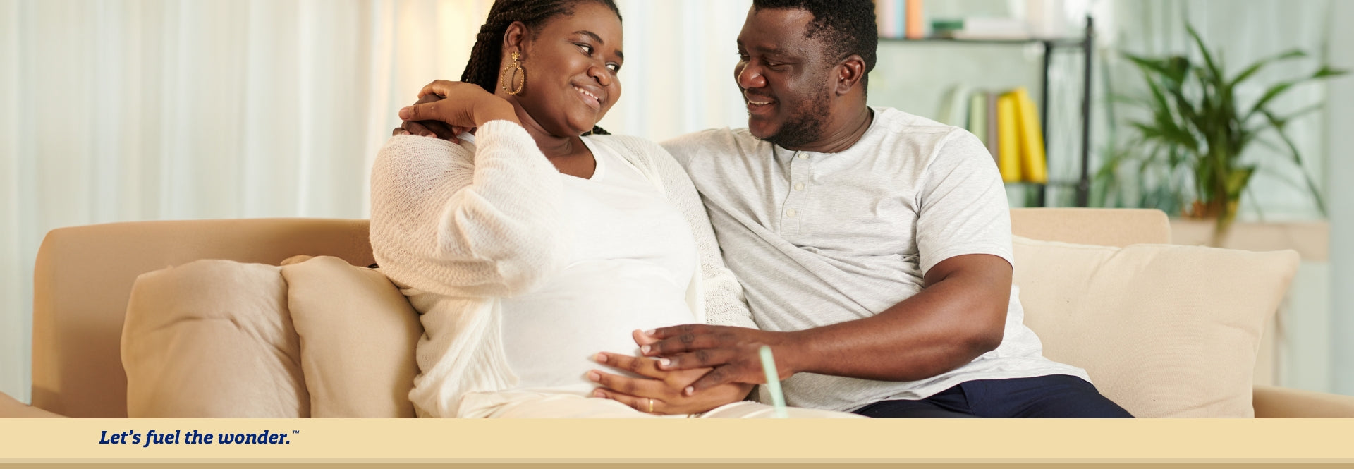 Pregnant mother sitting on couch hold pregnant belly in left hand. Father is sitting to pregnant mother's right side while glaring and smiling at pregnant mother. Father's right arm is on pregnant mother's right shoulder.