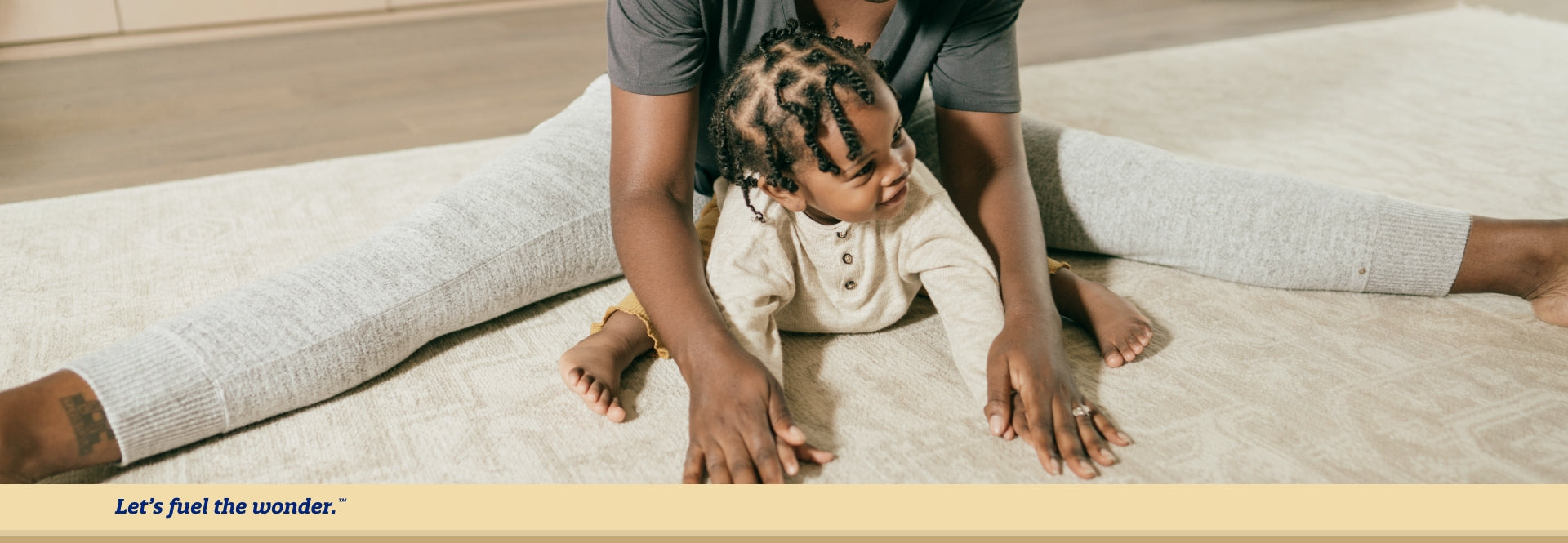 baby sitting forward on ground with arms and legs out while parent with legs spread out on floor sits behind baby holding baby's hands for support