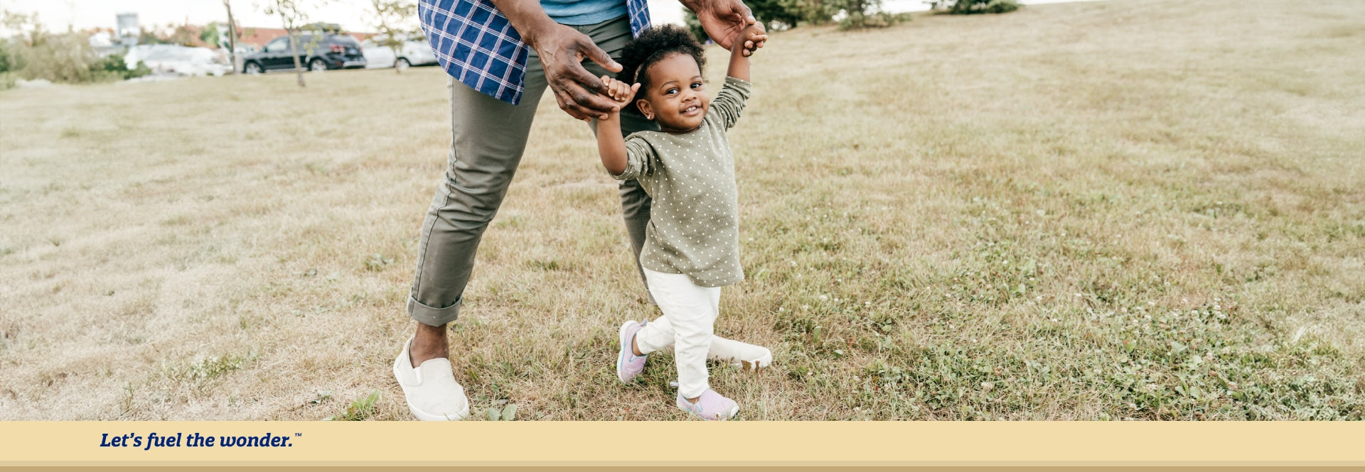 Older toddler outside walking on grass with arms held up by out of frame father figure.