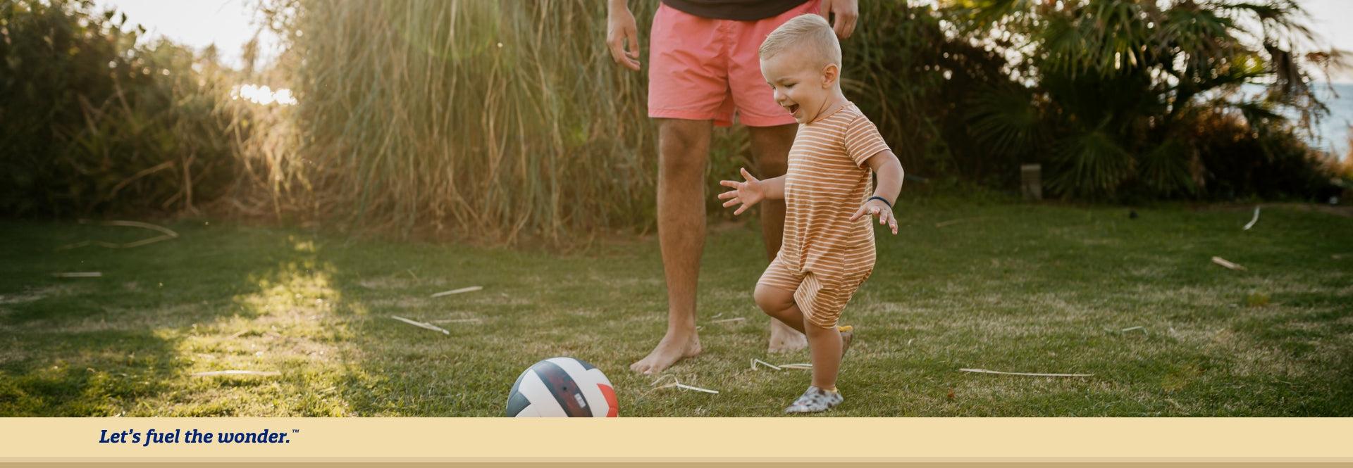 Older toddler outside running towards soccer ball preparing to kick it. Father is standing to the right of older toddler with only hands in legs visible in frame.
