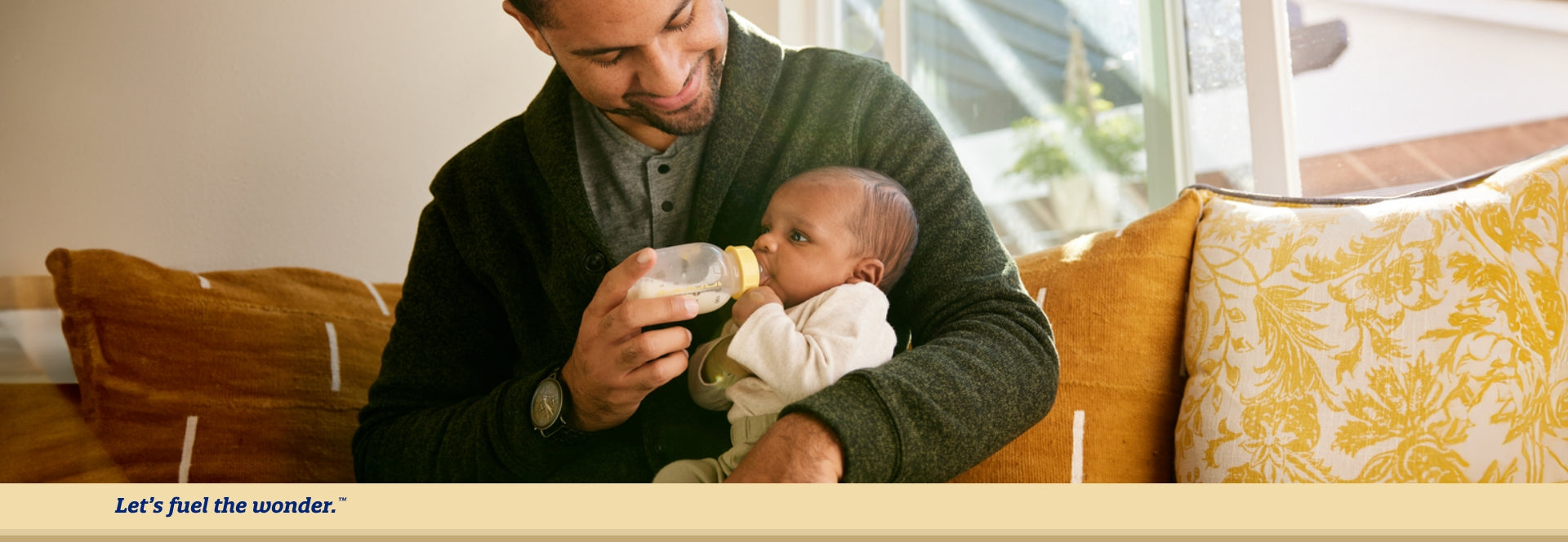 Baby sitting down on father's lap and held with father's left arm being fed from a bottle held by father's right hand. Father is smiling down at feeding baby.