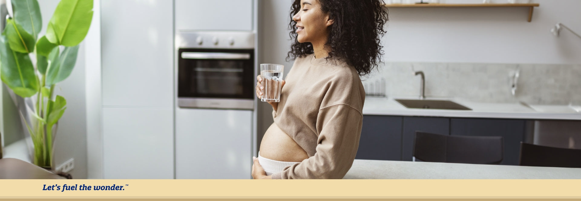 "Pregnant woman standing in kitchen with eyes closed, holding a  glass of water in right hand while holding pregnant belly with left  hand"