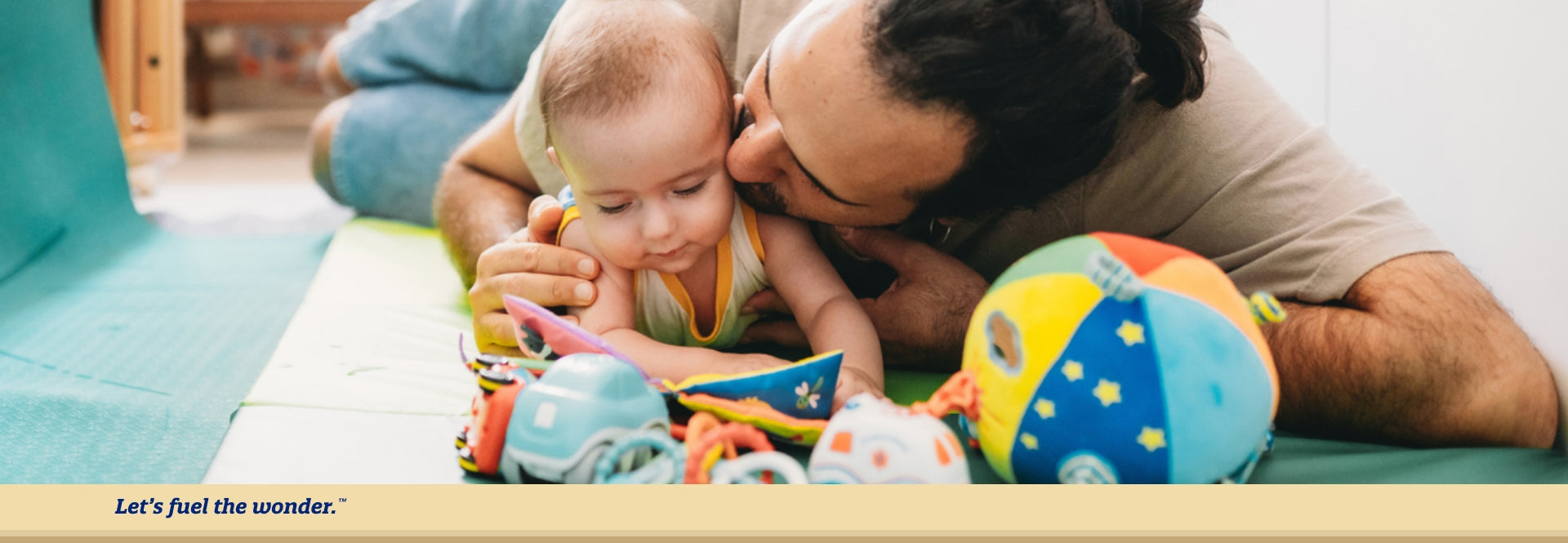 Baby laying on stomach on the ground while looking down at a toy book while father is laying down to their left with right hand on baby's right shoulder. Father is kissing baby on baby's left cheek.