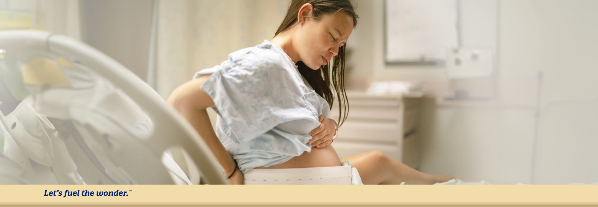 Pregnant parent sitting up in hospital bed displaying some discomfort while supporting their back with their right hand and holding their belly with their left hand
