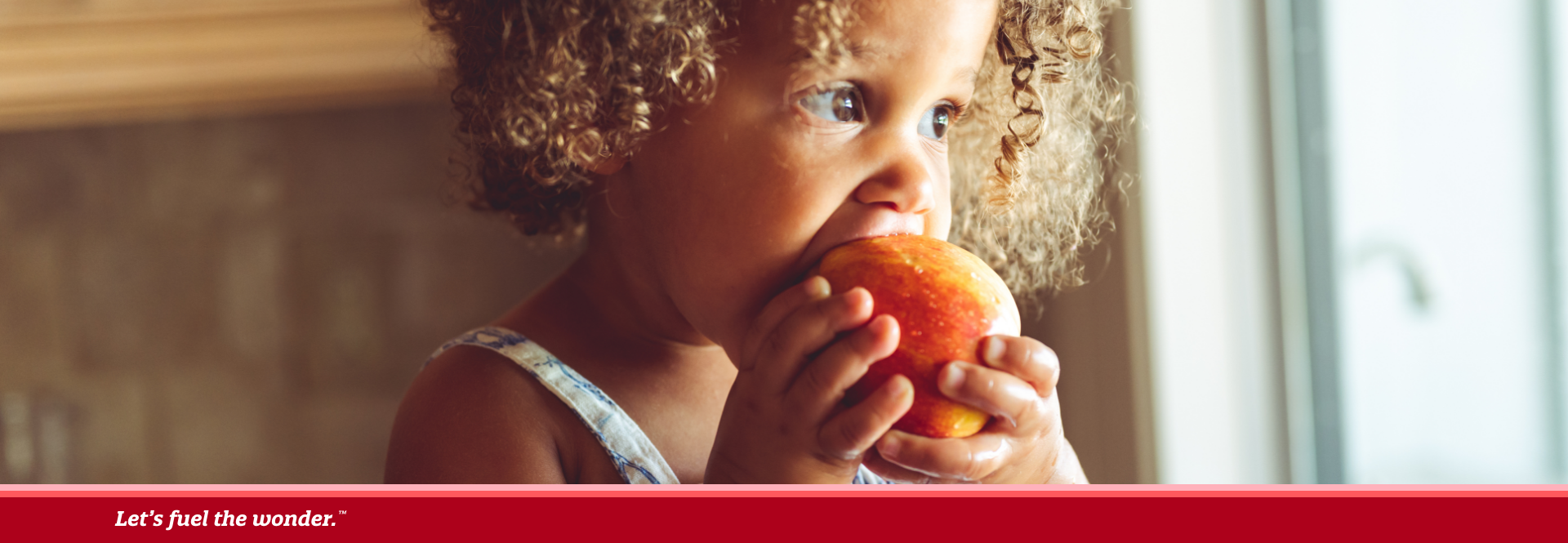 Young girl with curly hair eating an apple while standing near a window in a kitchen.