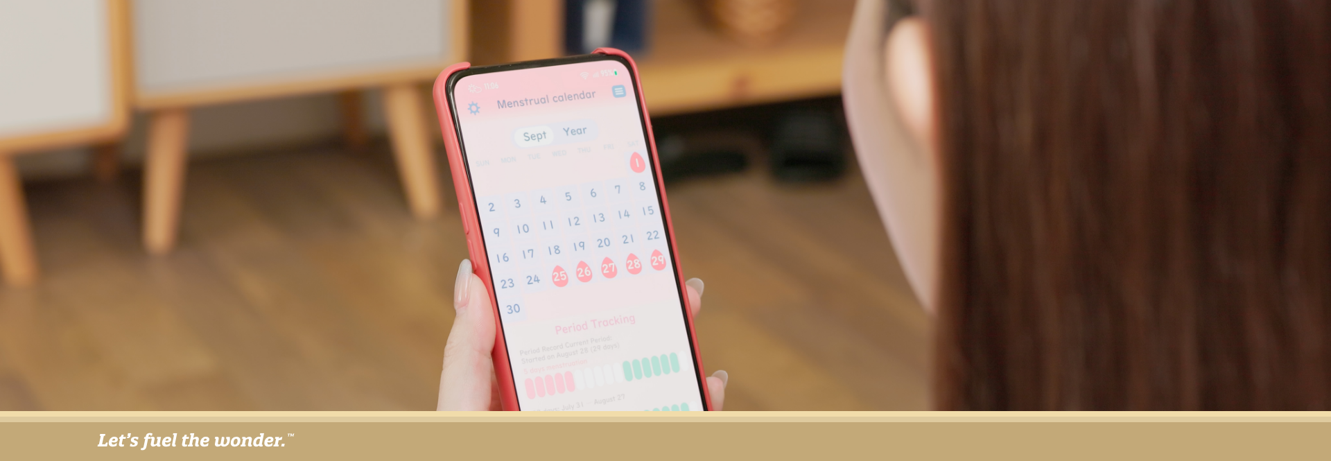 Woman holding a smartphone displaying a menstrual calendar app, viewed from behind as she sits indoors.