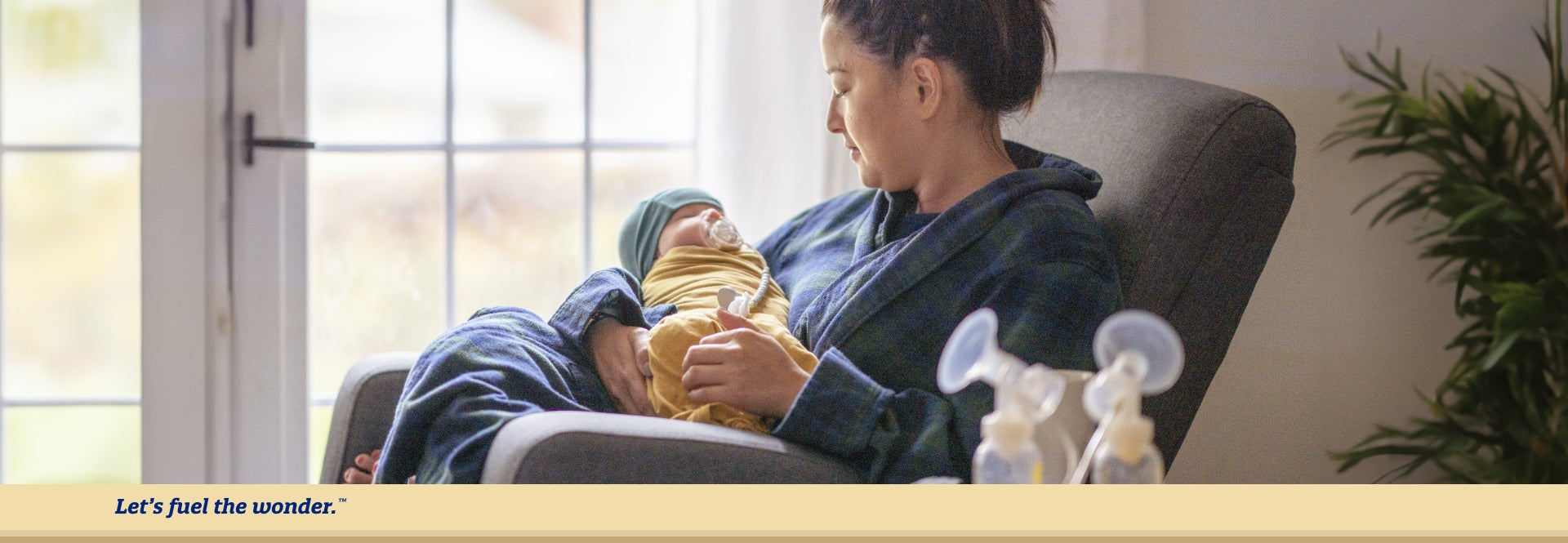 baby asleep and swaddled while being held by parent sitting in a chair with breast pumps resting on a table in the foreground