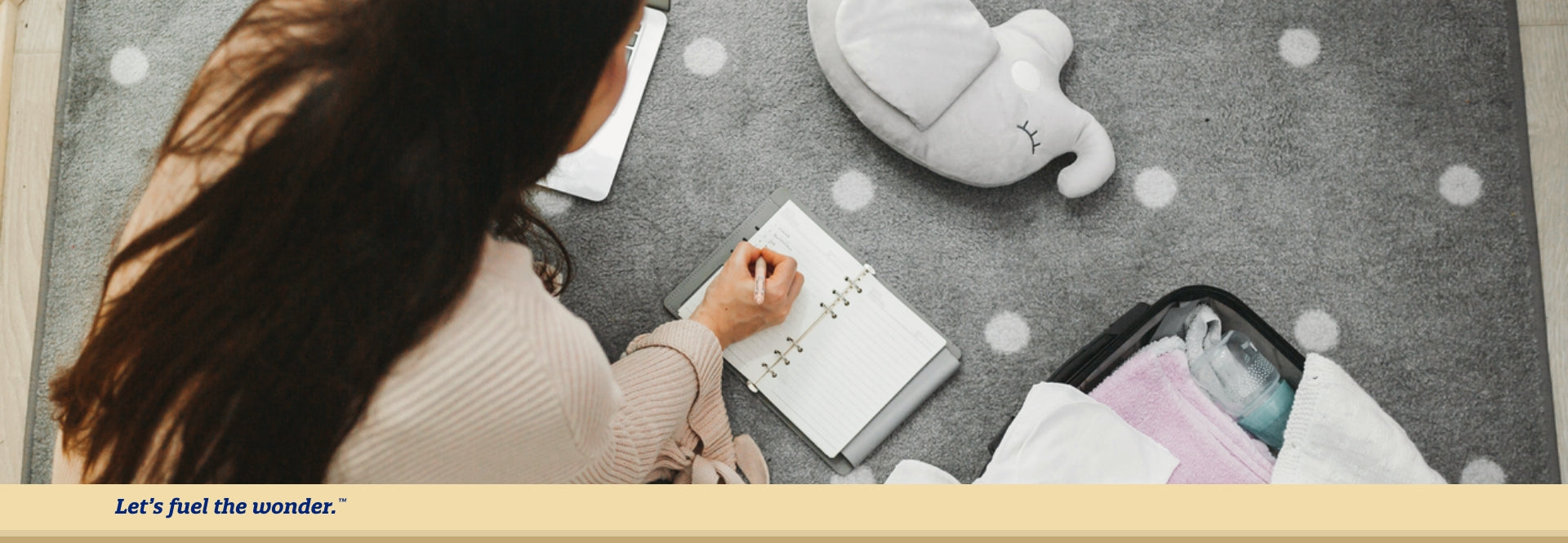 Woman sitting on ground writing in a notebook. Baby toys and supplies are scattered on ground around woman