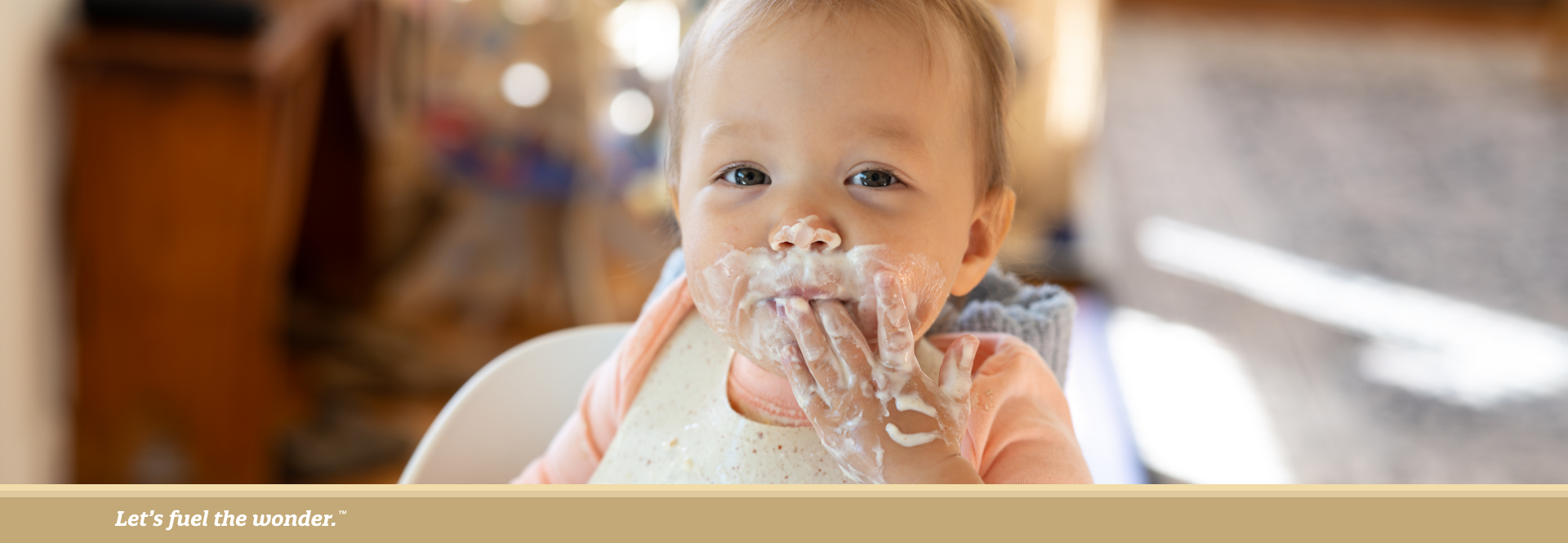 Baby sitting in a high chair with food all over their face and hands, wearing a bib and smiling while eating messily