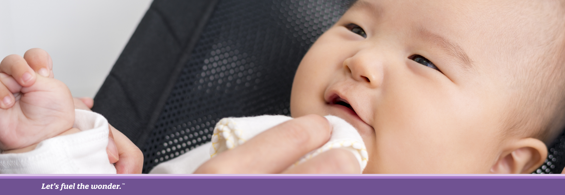 Smiling baby lying in a black mesh chair while a caregiver wipes their mouth with a cloth.