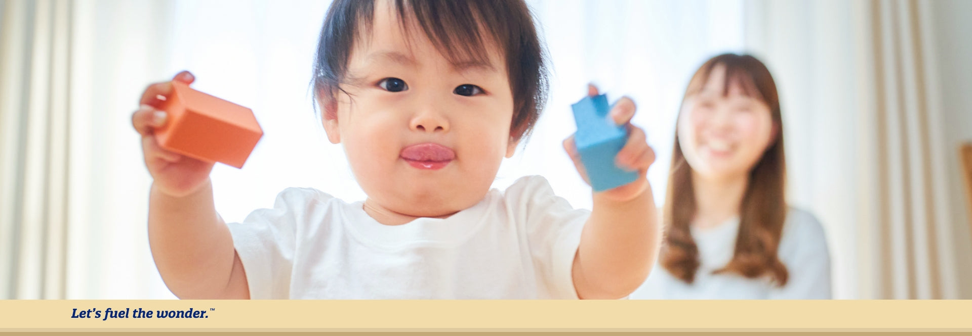 Young toddler licking their lips while walking towards camera with blocks in both of young toddlers hands. Out of focus mother is standing behind young toddler smiling at them