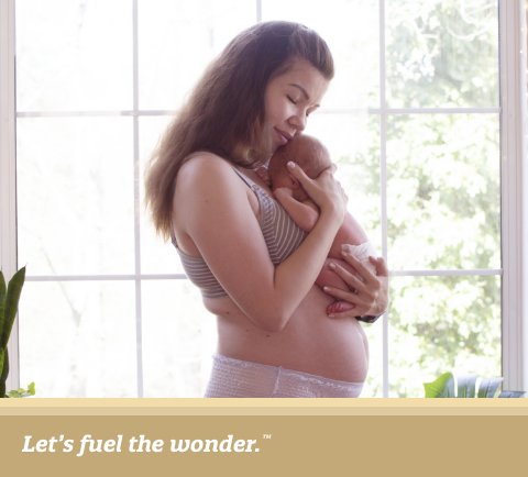 New mother in underwear gently holding her newborn baby near a large sunlit window.