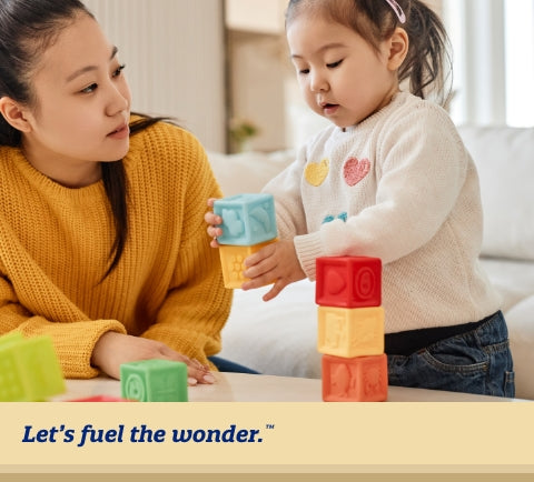 Older toddler standing in living room playing with building blocks on table while mother sitting and leaning their arm on table, staring at older toddler.