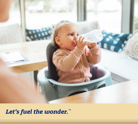 Young toddler sitting in high chair looking off to their left side as they are holding up and drinking from milk bottle