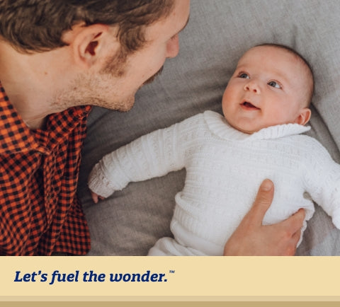 Baby laying on their back on a large bed with arms stretched out, staring up at baby's father as they look down towards baby and support baby's side with their right hand