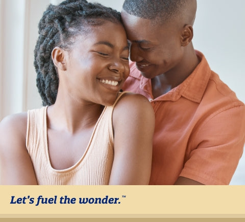 Smiling partner with eyes closed standing next to window as other smiling parent with eyes closed is hugging them from behind