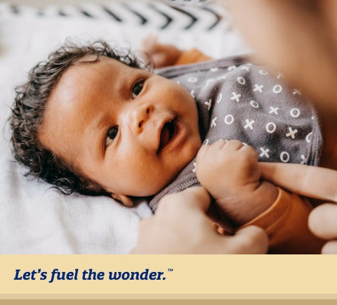Smiling baby looking up at parent while laying down on bed and grasping at parent's finger