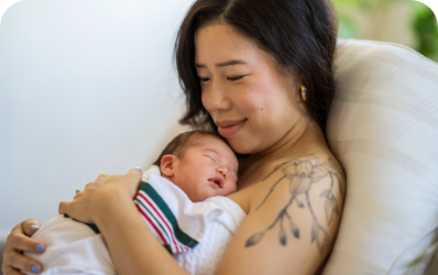 Mother cuddling her sleeping newborn, smiling gently while 
seated on a couch.