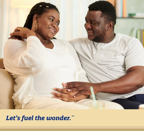 Pregnant mother sitting on couch hold pregnant belly in left hand. Father is sitting to pregnant mother's right side while glaring and smiling at pregnant mother. Father's right arm is on pregnant mother's right shoulder.