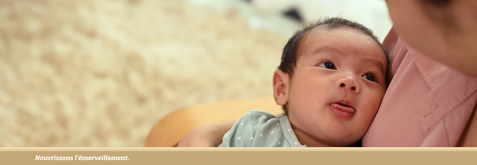 Newborn baby in patterned pyjamas sticks out their tongue 
while lying in a woman's arms. - FR