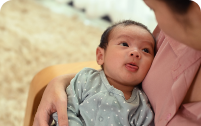 Newborn baby in patterned pyjamas sticks out their tongue 
while lying in a woman's arms.